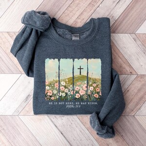 May include: A dark gray sweatshirt with a floral design and a cross on a hill. The text "He is not here. He has risen.  Matt. 28:6" is printed on the sweatshirt.