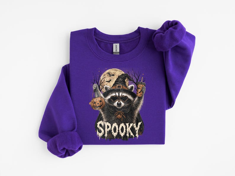 May include: A purple long-sleeve sweatshirt featuring a graphic of a raccoon wearing a witch hat and cape, holding a jack-o'-lantern and a drink. The background shows a full moon and bats, with the word SPOOKY printed in white distressed lettering.