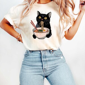 May include: A white t-shirt with a black cat eating ramen noodles. The cat is holding chopsticks in its paws and has a bowl of ramen in front of it. The ramen has a boiled egg and green onions.