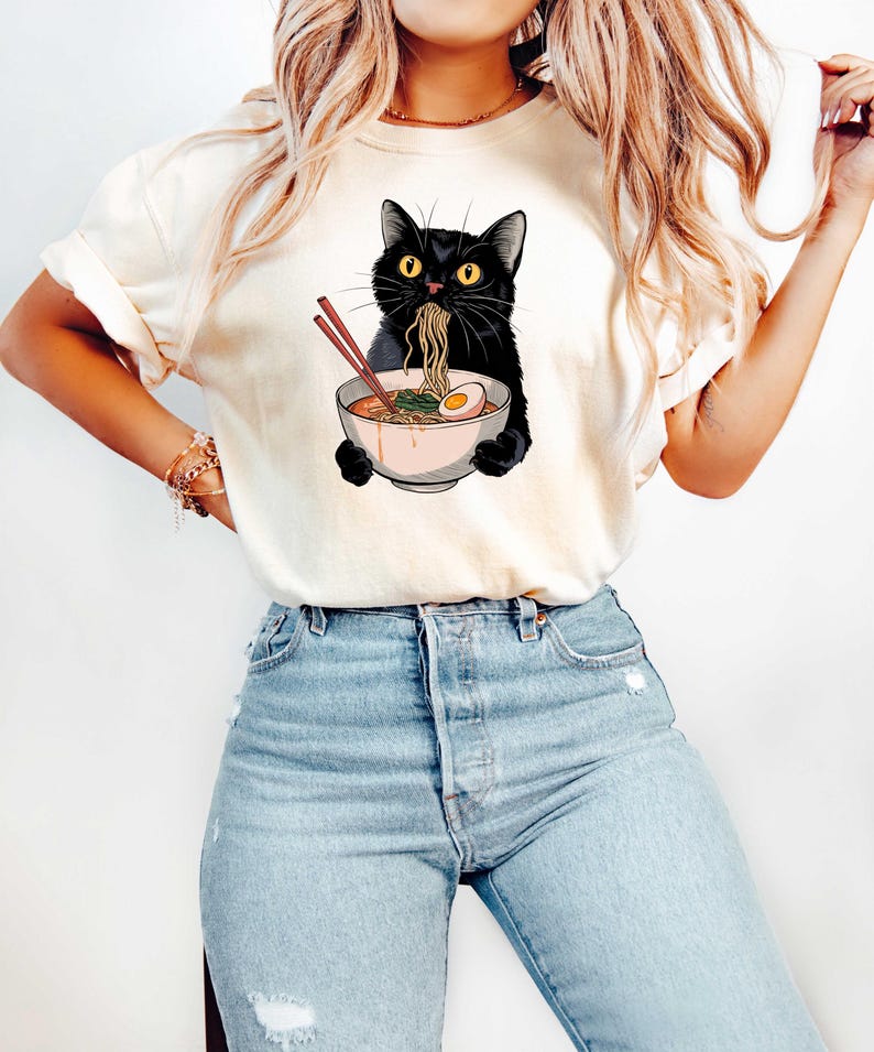 May include: A white t-shirt with a black cat eating ramen noodles. The cat is holding chopsticks in its paws and has a bowl of ramen in front of it. The ramen has a boiled egg and green onions.