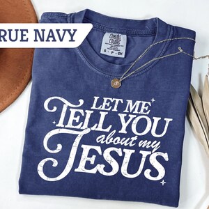 Puede incluir: Camiseta azul marino con el texto blanco "LET ME TELL YOU about my JESUS". La camiseta tiene cuello redondo y mangas cortas. Un collar dorado con un colgante est&aacute; sobre la camiseta.