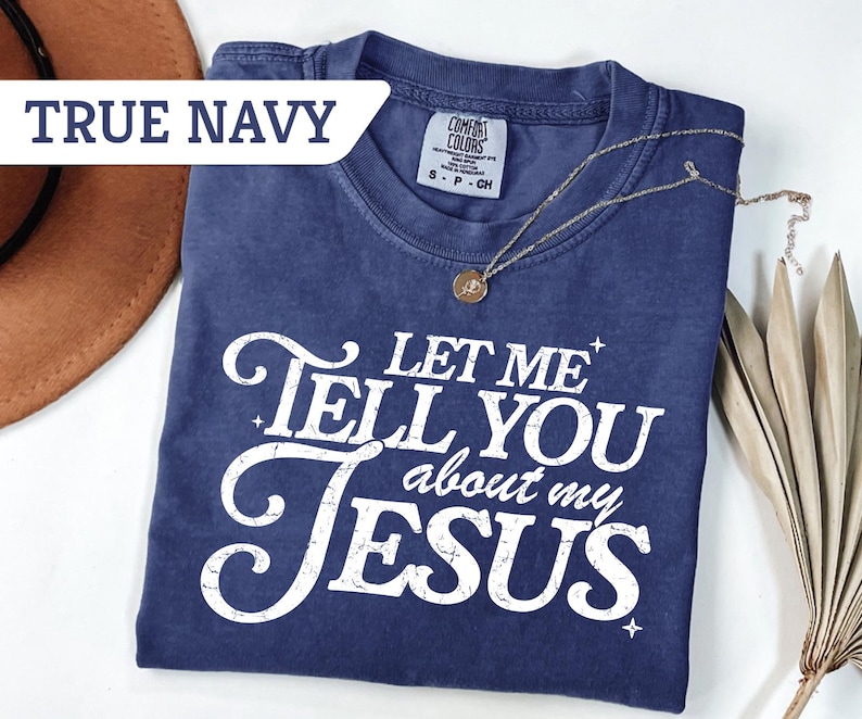 Puede incluir: Camiseta azul marino con el texto blanco "LET ME TELL YOU about my JESUS". La camiseta tiene cuello redondo y mangas cortas. Un collar dorado con un colgante est&aacute; sobre la camiseta.