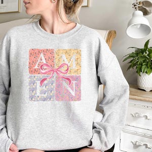 K&ouml;nnte beinhalten: Ein grauer Pullover mit einem Blumendesign, das "AMEN" mit einer rosa Schleife in der Mitte buchstabiert.