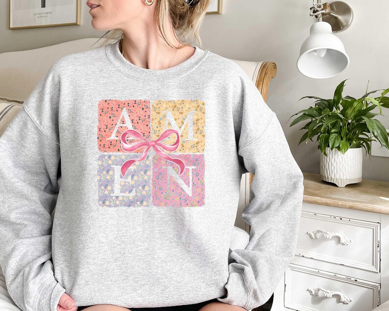 K&ouml;nnte beinhalten: Ein grauer Pullover mit einem Blumendesign, das "AMEN" mit einer rosa Schleife in der Mitte buchstabiert.