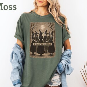 K&ouml;nnte beinhalten: Moosgr&uuml;nes T-Shirt mit einer Illustration im Vintage-Stil von vier Hexen in schwarzen Kleidern und H&uuml;ten, die in einem Wald unter einem Vollmond stehen. Der Text "GIRLS WILL BE GIRLS" ist darunter zu sehen.