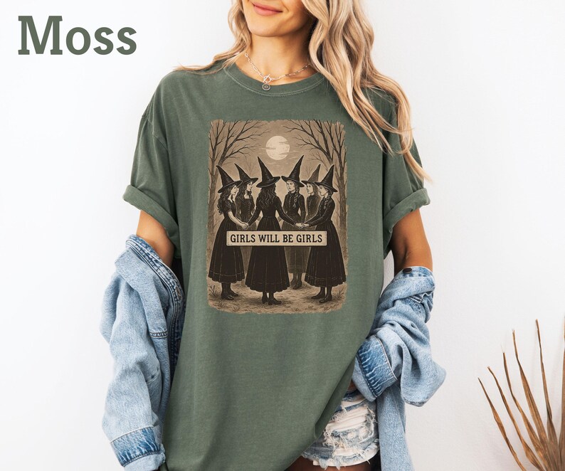 K&ouml;nnte beinhalten: Moosgr&uuml;nes T-Shirt mit einer Illustration im Vintage-Stil von vier Hexen in schwarzen Kleidern und H&uuml;ten, die in einem Wald unter einem Vollmond stehen. Der Text "GIRLS WILL BE GIRLS" ist darunter zu sehen.