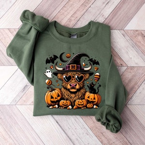 May include: A green sweatshirt with a cartoon highland cow wearing a witch hat and sunglasses. The cow is surrounded by pumpkins, candy corn, and bats.