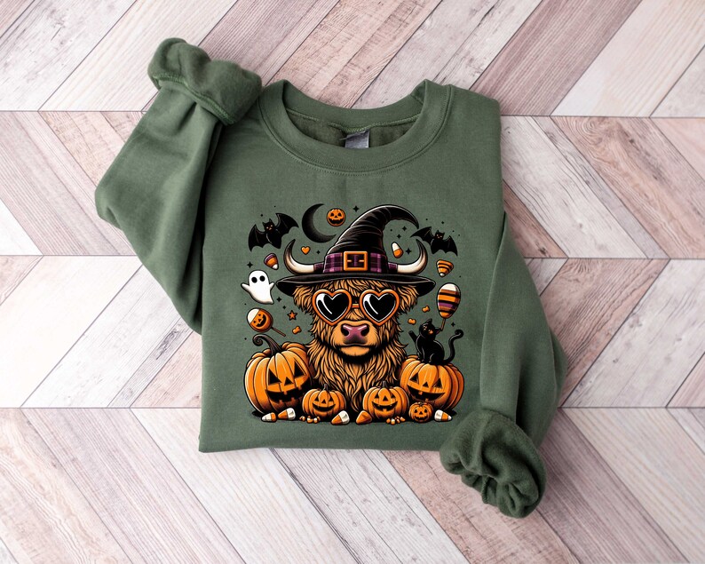 May include: A green sweatshirt with a cartoon highland cow wearing a witch hat and sunglasses. The cow is surrounded by pumpkins, candy corn, and bats.