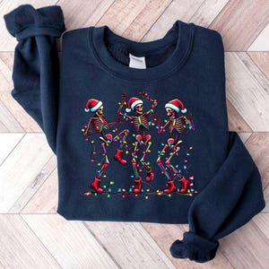 May include: A navy blue sweatshirt with a Christmas-themed design featuring three skeletons wearing Santa hats and dancing with colorful lights.
