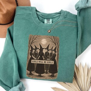 K&ouml;nnte beinhalten: Ein teal Comfort Colors Sweatshirt mit einem sepiafarbenen Aufdruck von sechs Frauen mit Hexenh&uuml;ten, die sich an den H&auml;nden halten, mit dem Text "GIRLS WILL BE GIRLS". Das Design ist vor einem Vollmond und kahlen B&auml;umen platziert. Eine goldene Halskette ist sichtbar.