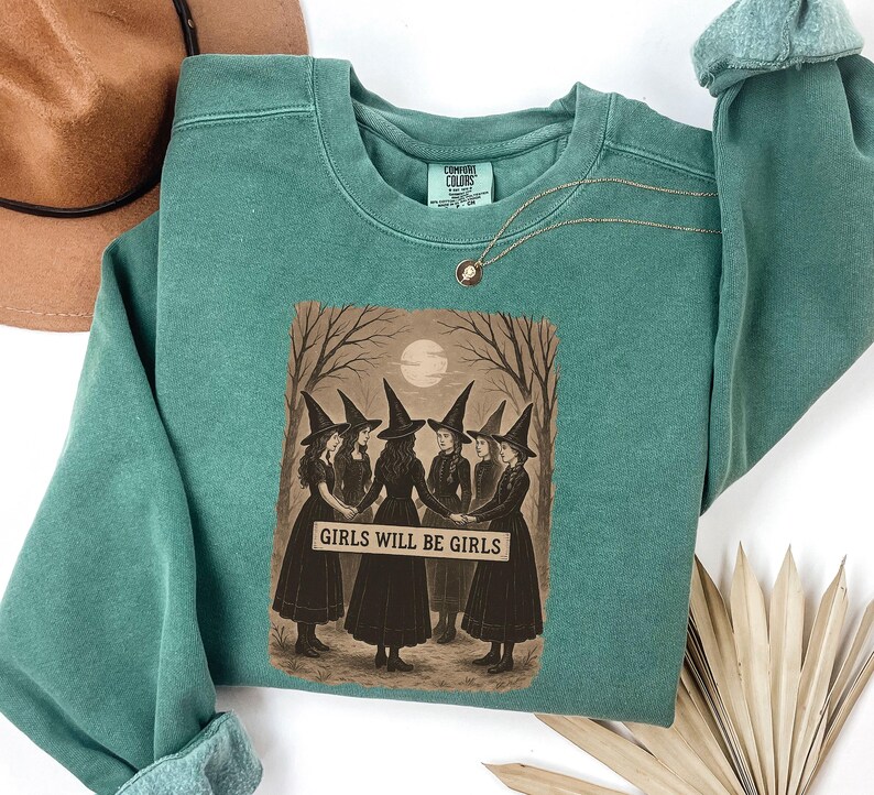 K&ouml;nnte beinhalten: Ein teal Comfort Colors Sweatshirt mit einem sepiafarbenen Aufdruck von sechs Frauen mit Hexenh&uuml;ten, die sich an den H&auml;nden halten, mit dem Text "GIRLS WILL BE GIRLS". Das Design ist vor einem Vollmond und kahlen B&auml;umen platziert. Eine goldene Halskette ist sichtbar.