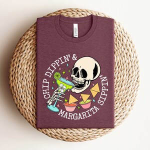 Pu&ograve; includere: Una maglietta color marrone con una grafica bianca di uno scheletro che beve una margarita e il testo "Chip Dippin' & Sippin' Margarita".