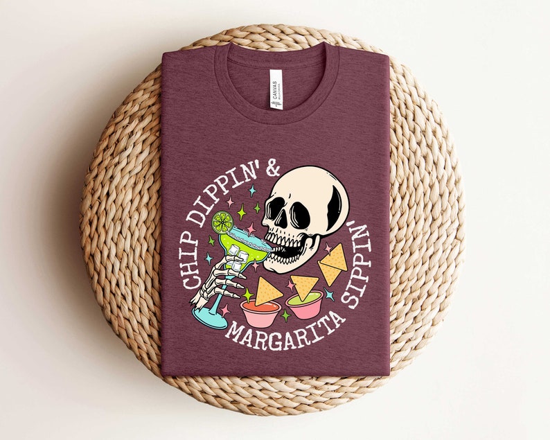 Pu&ograve; includere: Una maglietta color marrone con una grafica bianca di uno scheletro che beve una margarita e il testo "Chip Dippin' & Sippin' Margarita".