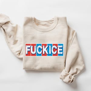 以下が含まれることがあります： 赤、白、青のデザインで「FUCKICE」の文字がプリントされたベージュ色のスウェットシャツ。スウェットシャツは折りたたまれ、袖は丸められています。グラフィックは言葉遊びです。