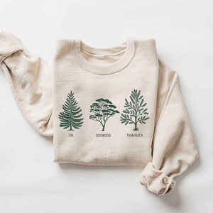 Pu&ograve; includere: Una felpa beige con una grafica di tre alberi, etichettati "Fir", "Dogwood" e "Tamarack".
