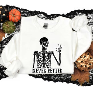 May include: White sweatshirt with a black skeleton graphic and the text "Never Better". The skeleton is making an "okay" gesture with its hand. The sweatshirt is surrounded by black netting and fall-themed decorations.