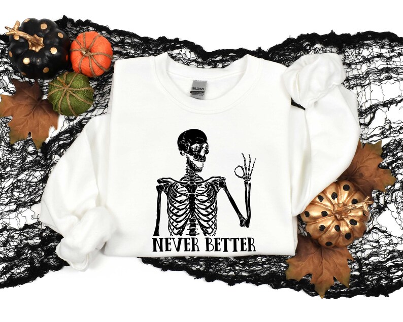 May include: White sweatshirt with a black skeleton graphic and the text "Never Better". The skeleton is making an "okay" gesture with its hand. The sweatshirt is surrounded by black netting and fall-themed decorations.