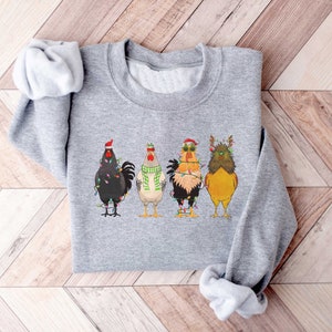 May include: A gray sweatshirt with a festive design featuring four roosters wearing Christmas hats and lights. The roosters are standing in a row, with the one on the left wearing a Santa hat, the one in the middle wearing a green scarf, the one on the right wearing sunglasses, and the one on the far right wearing reindeer antlers.