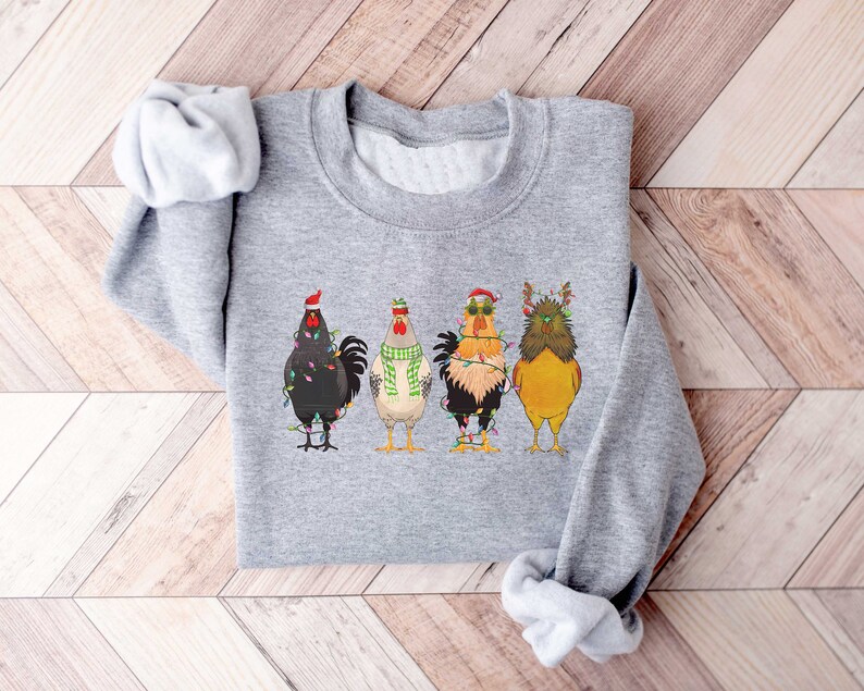 May include: A gray sweatshirt with a festive design featuring four roosters wearing Christmas hats and lights. The roosters are standing in a row, with the one on the left wearing a Santa hat, the one in the middle wearing a green scarf, the one on the right wearing sunglasses, and the one on the far right wearing reindeer antlers.