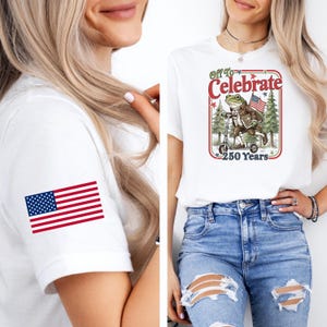 May include: White t-shirt with a graphic of a frog riding a scooter, holding an American flag, and the text "Off to Celebrate 250 Years." The shirt also features a small American flag on the sleeve. The person is wearing blue jeans.