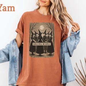 K&ouml;nnte beinhalten: Ein yam-farbenes T-Shirt mit einem Vintage-Grafik von f&uuml;nf Hexen in schwarzen Kleidern und H&uuml;ten, die sich unter einem Vollmond an den H&auml;nden halten. Der Text "GIRLS WILL BE GIRLS" ist unter dem Bild aufgedruckt. Das Shirt hat hochgekrempelte &Auml;rmel.