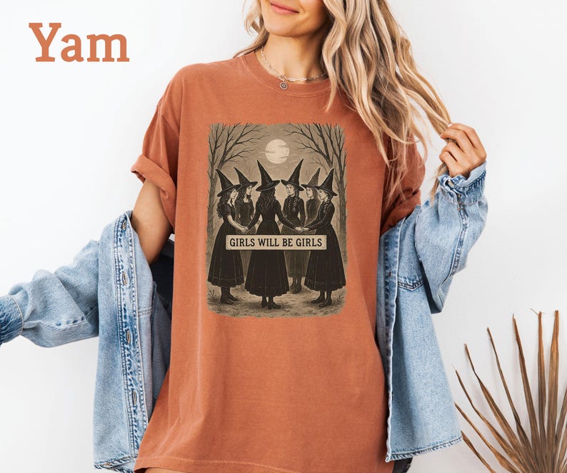 K&ouml;nnte beinhalten: Ein yam-farbenes T-Shirt mit einem Vintage-Grafik von f&uuml;nf Hexen in schwarzen Kleidern und H&uuml;ten, die sich unter einem Vollmond an den H&auml;nden halten. Der Text "GIRLS WILL BE GIRLS" ist unter dem Bild aufgedruckt. Das Shirt hat hochgekrempelte &Auml;rmel.