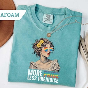 May include: Seafoam green t-shirt with a graphic of a woman wearing rainbow heart-shaped sunglasses and yellow flowers in her hair. The text "MORE PRIDE LESS PREJUDICE" is printed below the graphic. A gold necklace is draped on the shirt.