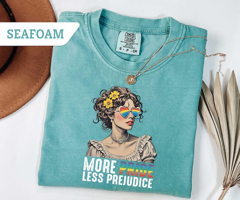 May include: Seafoam green t-shirt with a graphic of a woman wearing rainbow heart-shaped sunglasses and yellow flowers in her hair. The text "MORE PRIDE LESS PREJUDICE" is printed below the graphic. A gold necklace is draped on the shirt.