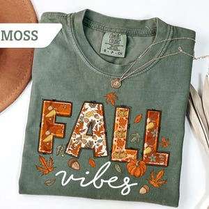 May include: A moss green t-shirt with the words "FALL vibes" in a fall-themed design. The word "FALL" is in orange and brown with fall leaves and pumpkin details. A silver necklace is draped across the shirt.