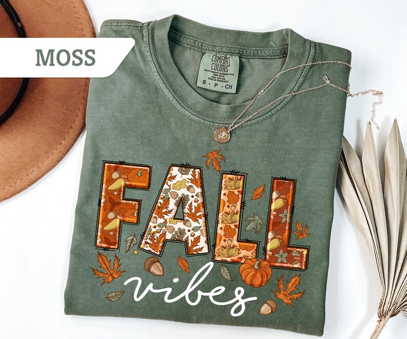 May include: A moss green t-shirt with the words "FALL vibes" in a fall-themed design. The word "FALL" is in orange and brown with fall leaves and pumpkin details. A silver necklace is draped across the shirt.