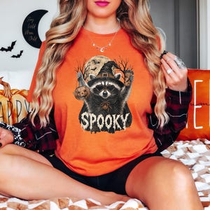 May include: An orange t-shirt featuring a graphic of a raccoon wearing a witch hat, holding a small pumpkin and a coffee cup. The design includes a full moon, bare tree silhouettes, and the word SPOOKY in a dripping, distressed white font.