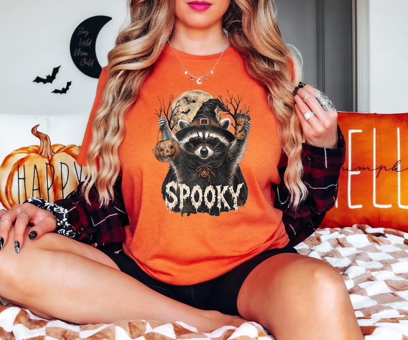 May include: An orange t-shirt featuring a graphic of a raccoon wearing a witch hat, holding a small pumpkin and a coffee cup. The design includes a full moon, bare tree silhouettes, and the word SPOOKY in a dripping, distressed white font.