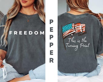 May include: Dark gray t-shirt with the word "FREEDOM" in white block letters on the front. The back features an American flag graphic with the text "This is the Turning Point."