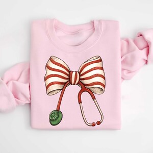 May include: A light pink sweatshirt featuring a whimsical illustration of a bow with red and white stripes and a red stethoscope. The stethoscope has a green earpiece. The sweatshirt is folded with the sleeves visible.