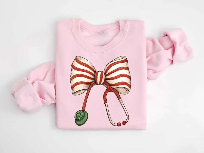 May include: A light pink sweatshirt featuring a whimsical illustration of a bow with red and white stripes and a red stethoscope. The stethoscope has a green earpiece. The sweatshirt is folded with the sleeves visible.