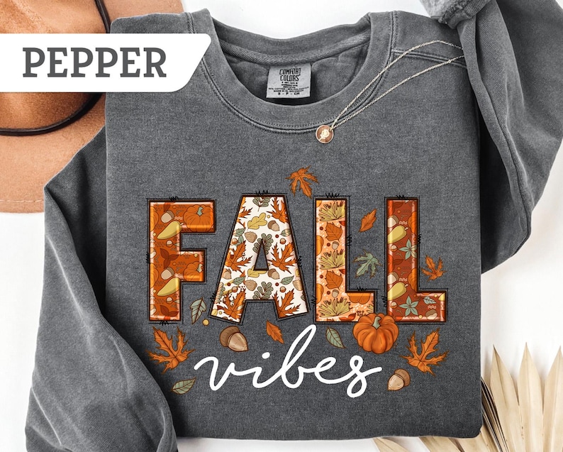May include: A dark gray sweatshirt with the words "FALL vibes" in a fall-themed design. The letters are filled with autumn patterns and surrounded by leaves, pumpkins, and acorns. The word "PEPPER" is in a white rectangle in the upper left corner.