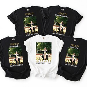 May include: Five t-shirts, black and white, with gold text and a photo. The shirts read "Proud Dad of a," "Proud Brother of a," "Proud Grandfather of a," and "Proud Sister of a." Each shirt has a photo of a person and the name "Katie Williams" with the year "2026."