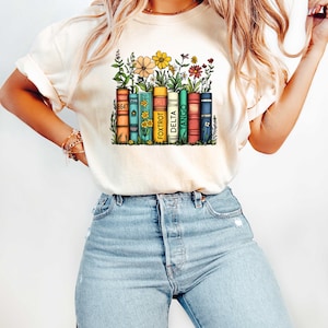 Peut inclure: Un t-shirt blanc avec un motif repr&eacute;sentant une pile de livres avec des fleurs qui poussent dessus. Les livres ont les mots "8647", "FOXTROT", "DELTA", "TANGO" imprim&eacute;s sur leur dos.