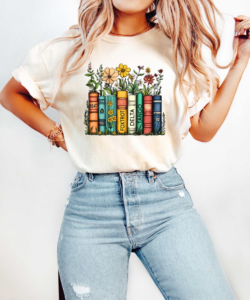 Peut inclure: Un t-shirt blanc avec un motif repr&eacute;sentant une pile de livres avec des fleurs qui poussent dessus. Les livres ont les mots "8647", "FOXTROT", "DELTA", "TANGO" imprim&eacute;s sur leur dos.