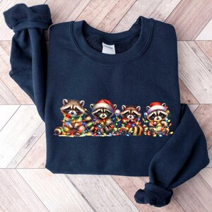 May include: A navy blue sweatshirt with a Christmas-themed design featuring four raccoons wearing Santa hats and holding strings of Christmas lights.