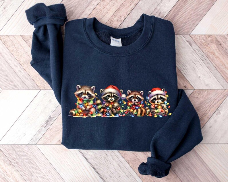 May include: A navy blue sweatshirt with a Christmas-themed design featuring four raccoons wearing Santa hats and holding strings of Christmas lights.