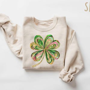 May include: A beige sweatshirt with a green, gold, and pink four-leaf clover design. The sweatshirt is called "SAND".