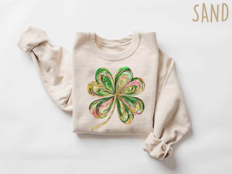 May include: A beige sweatshirt with a green, gold, and pink four-leaf clover design. The sweatshirt is called "SAND".