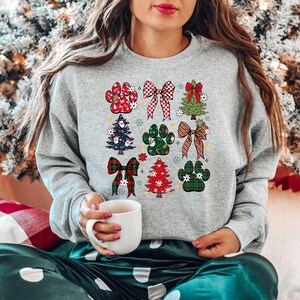 May include: A gray sweatshirt with a Christmas-themed design featuring paw prints, bows, and Christmas trees. The design is in red, green, and white.