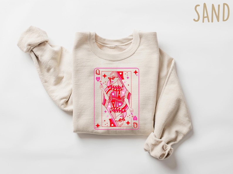 May include: A sand-colored sweatshirt with a pink and red Queen of Hearts playing card graphic. The graphic features a woman wearing sunglasses and a crown.