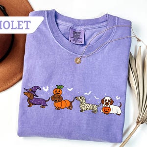 May include: Violet Comfort Colors t-shirt featuring four dachshunds in Halloween costumes: a witch, a pumpkin, a mummy, and a ghost. The shirt has a crew neck and a small tag on the collar. A gold necklace with a pendant is draped across the shirt.