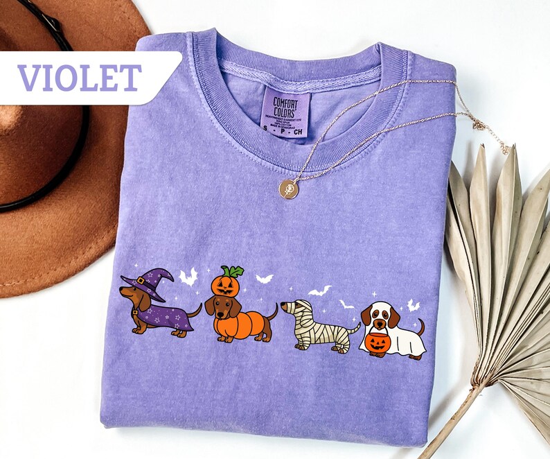 May include: Violet Comfort Colors t-shirt featuring four dachshunds in Halloween costumes: a witch, a pumpkin, a mummy, and a ghost. The shirt has a crew neck and a small tag on the collar. A gold necklace with a pendant is draped across the shirt.