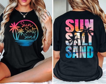 Può includere: T-shirt nero con grafica colorata. La parte anteriore presenta una palma e le parole "Sun Salt Sand" in un design circolare. La parte posteriore della maglietta ha le stesse parole impilate verticalmente con una scena di spiaggia e palme.