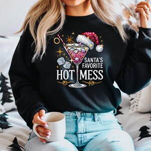 May include: A black sweatshirt with a glittery design of a martini glass with a Santa hat on top. The text "Santa's Favorite Hot Mess" is printed below the glass.