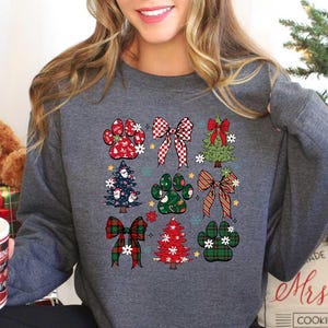 May include: A gray sweatshirt with a Christmas-themed design featuring paw prints, bows, and Christmas trees. The design is in red, green, and white.